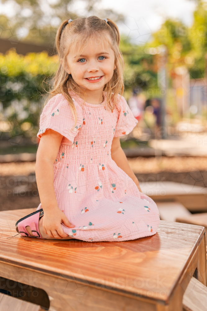 A happy little girl sitting on a wooden box outdoors at kindergarten - Australian Stock Image