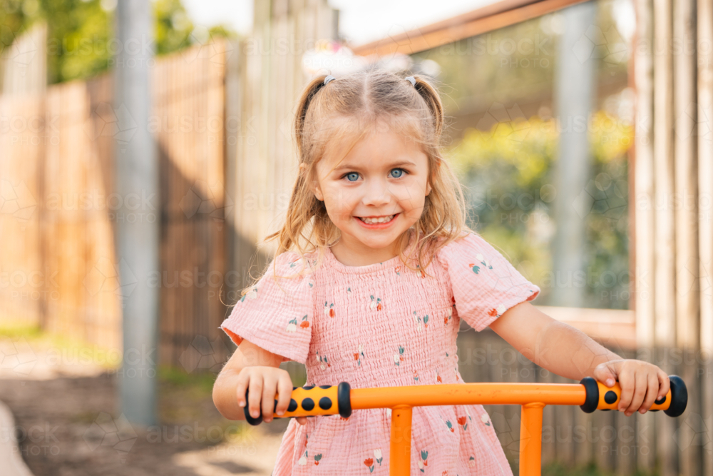 A happy preschool age girl riding orange trike at kindergarten - Australian Stock Image