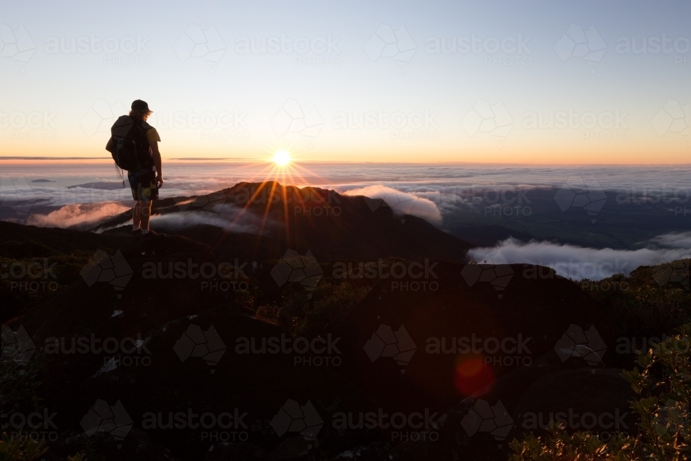 A hiker watches a beautiful sunrise on from the summit of a mountain above the clouds in Queensland. - Australian Stock Image