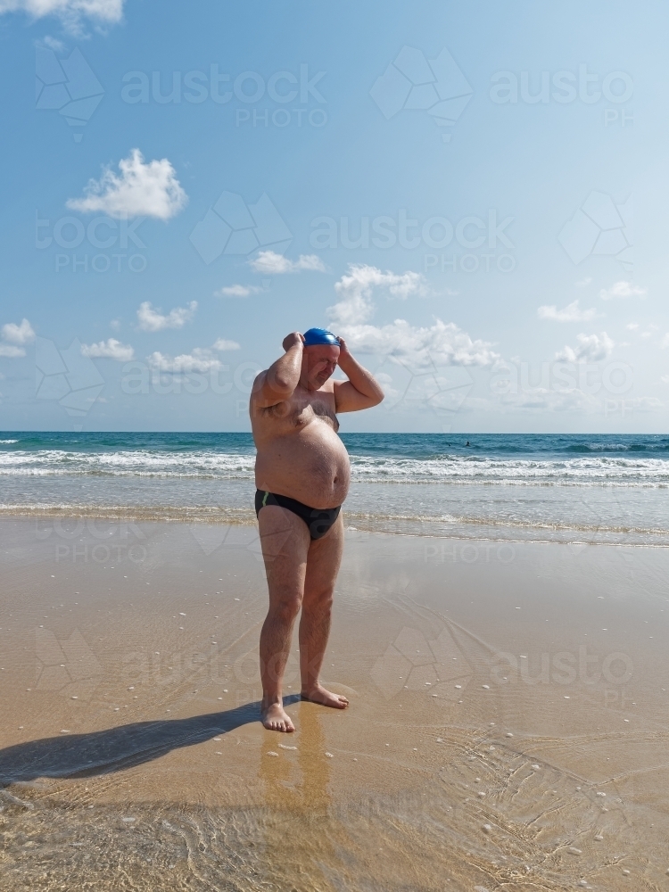 A large man putting on a swimming cap standing on an empty beach - Australian Stock Image