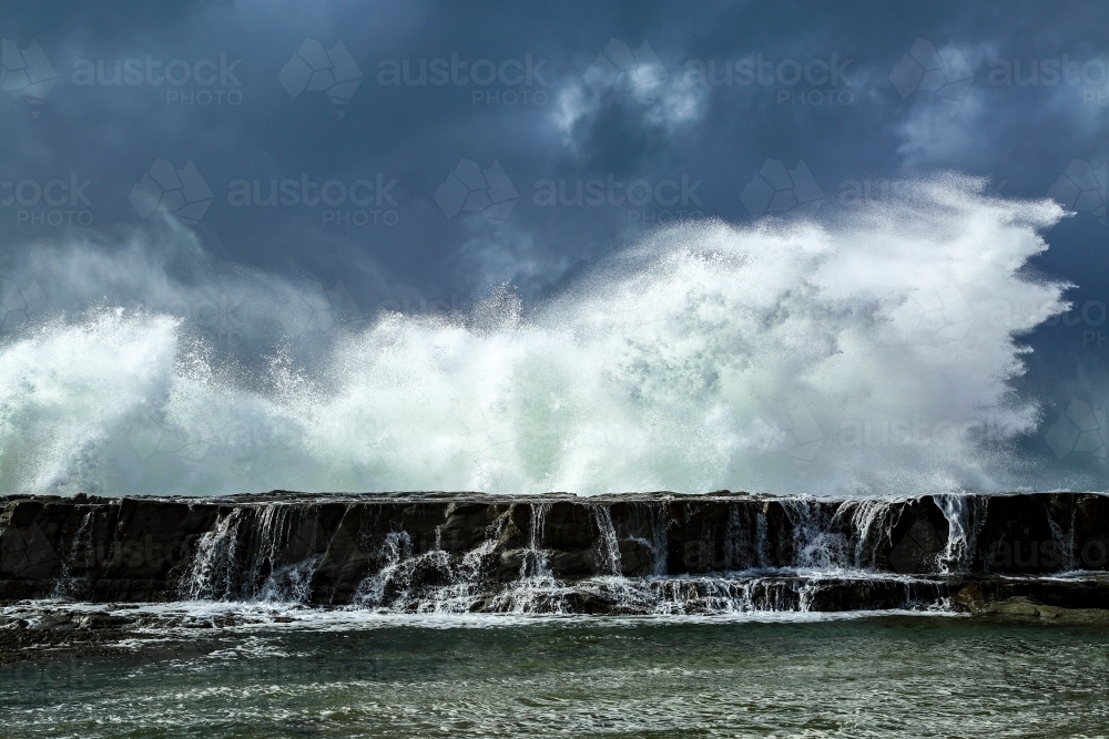 A large swell smashes against a rocky headland under a stormy sky in Iluka, New South Wales - Australian Stock Image