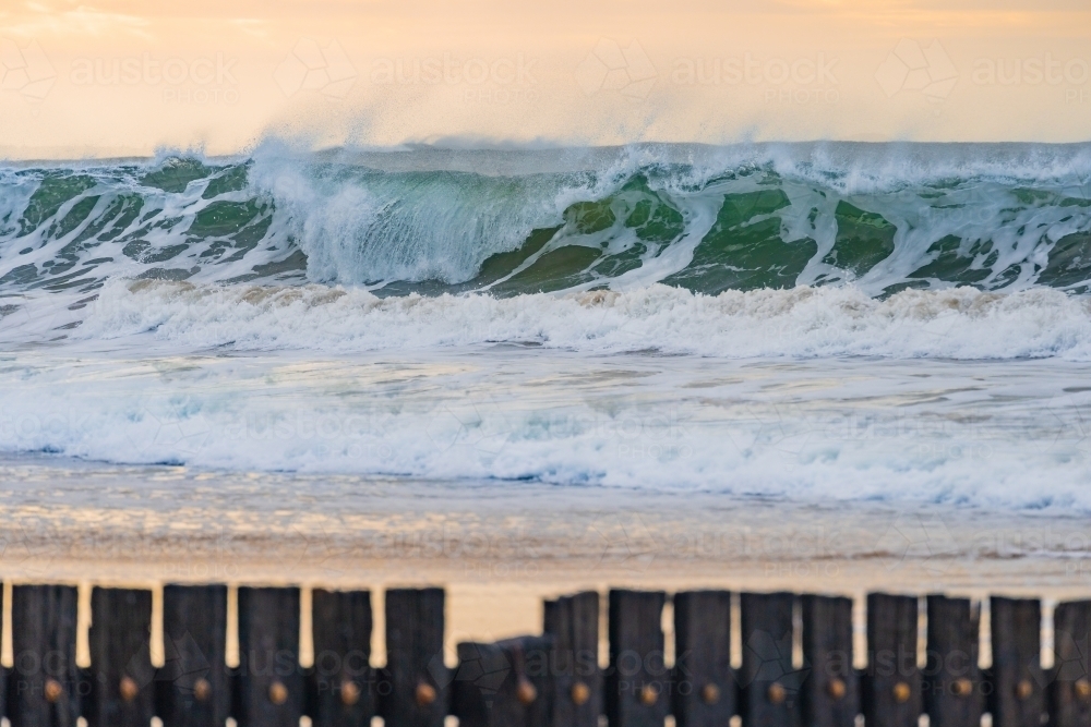 A large wave crashing with spray coming of the crest and a wooden groyne on the foreground - Australian Stock Image