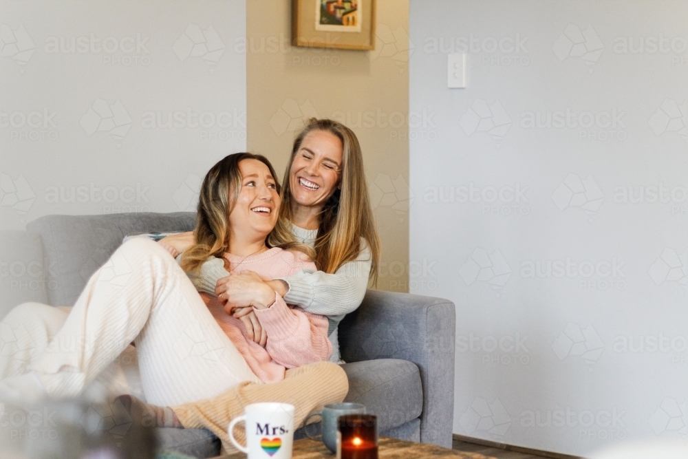 A lesbian couple laughing, talking and hugging on the couch - Australian Stock Image