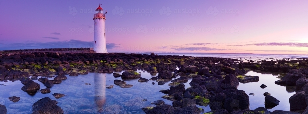 A lighthouse reflected in rock pools against a purple dawn sky - Australian Stock Image