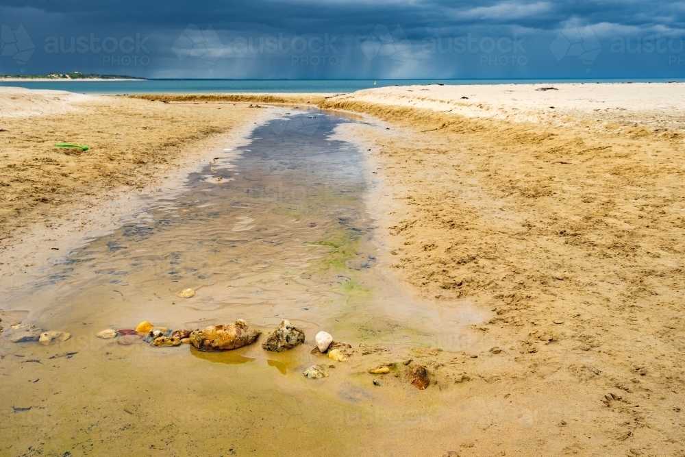 A line of rocks crossing a small creek crossing a sandy beach with rain falling overhead - Australian Stock Image