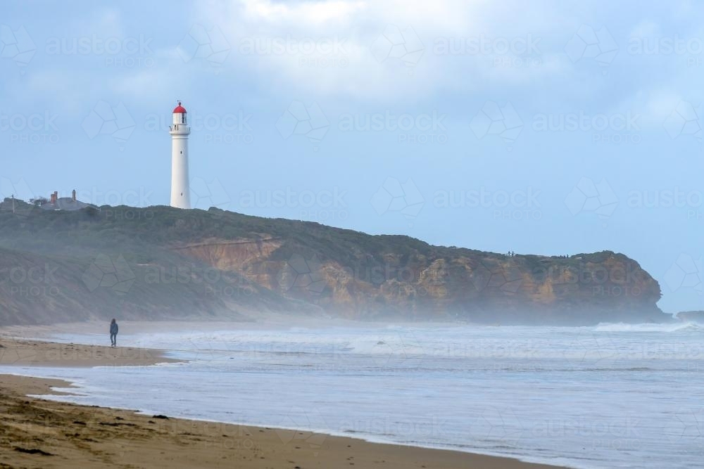 A lone figure on a beach below cliffs and a lighthouse - Australian Stock Image