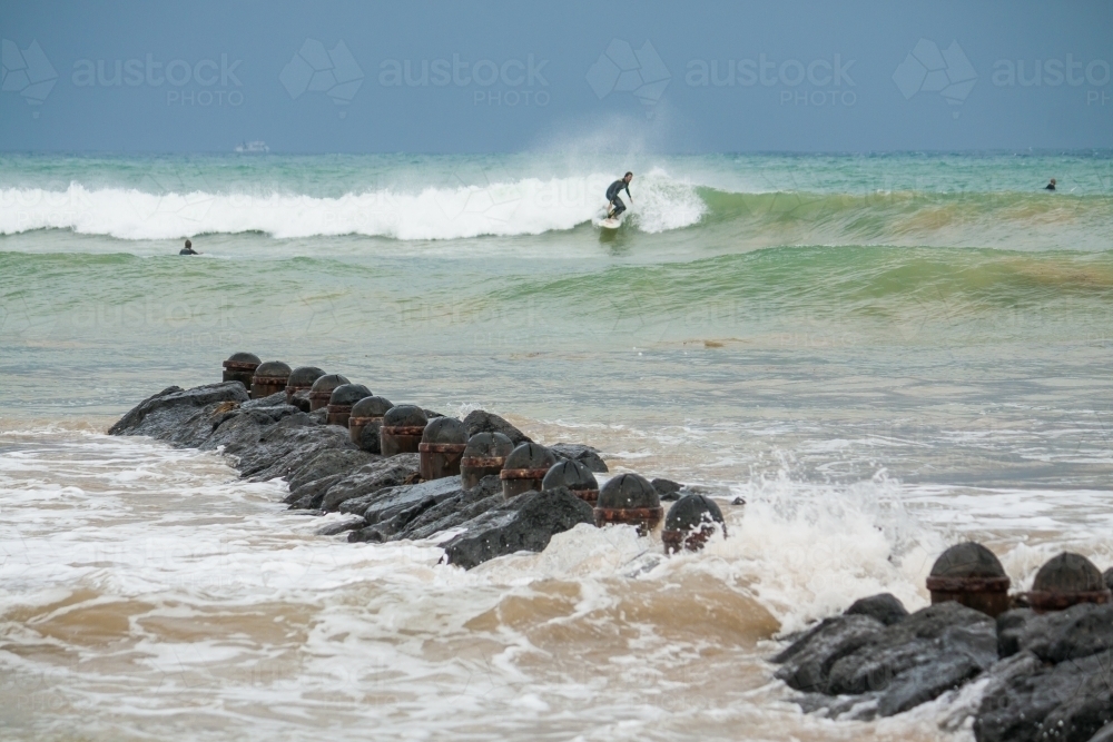 A lone surfer rides a wave in towards a rocky groyne on a stormy day - Australian Stock Image