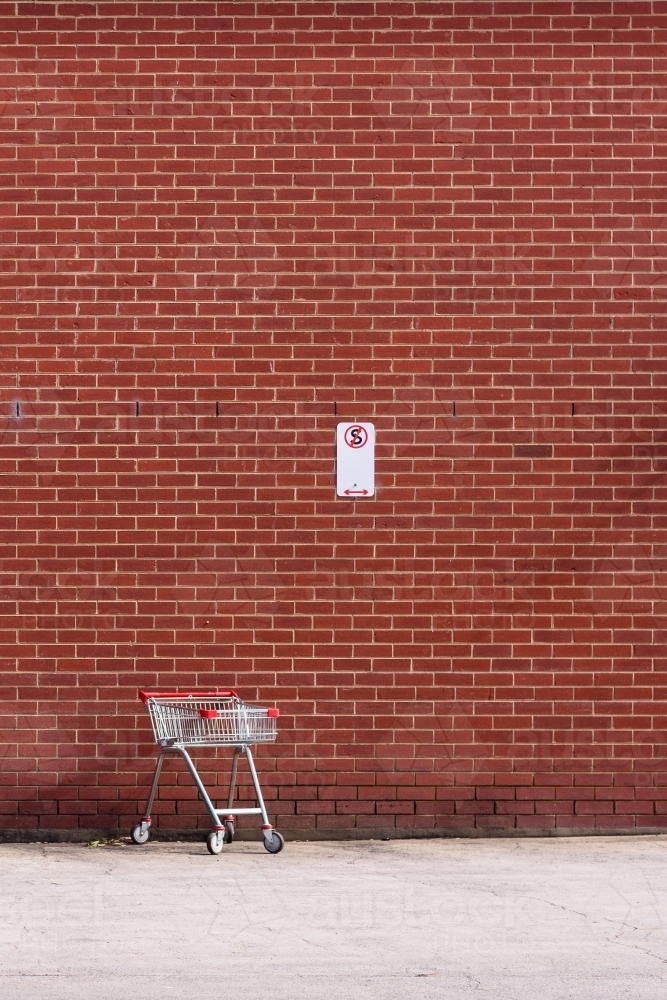 A lonely shopping trolley stays in a non-stopping open space, against a red brick wall. - Australian Stock Image
