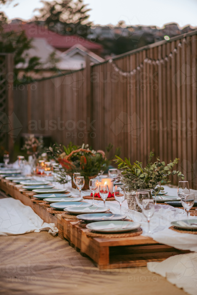 A long wooden table adorned with elegant tableware and glassware is set for an outdoor dinner - Australian Stock Image