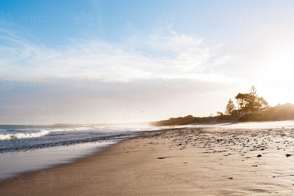 A low view of hundreds of pebbles spread wet sand at the shoreline of a beach, as the sun sets. - Australian Stock Image