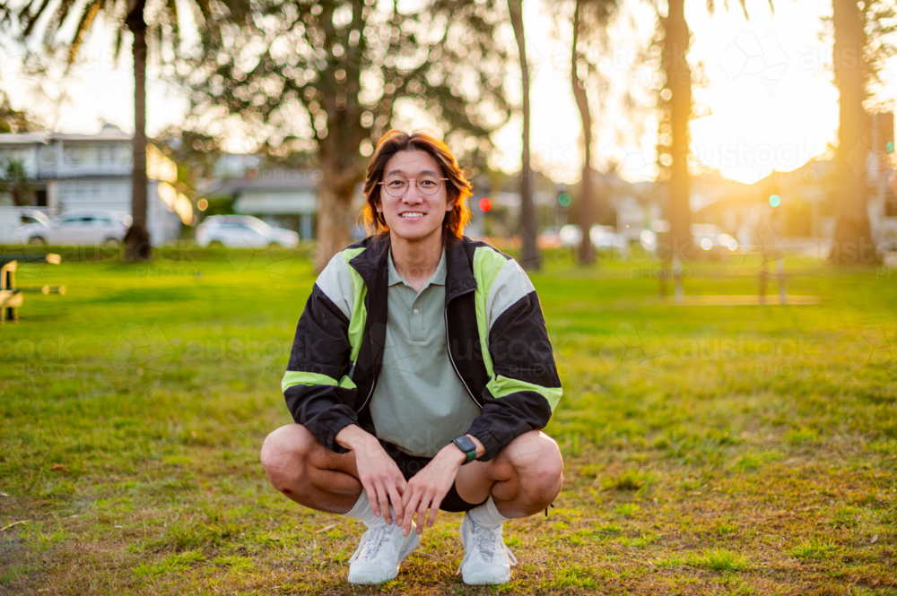 A male in athletic wear squats on grass, smiling as the sun sets behind the trees - Australian Stock Image