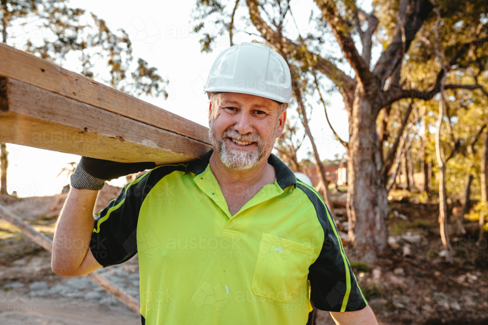 A man carrying a stack of lumber on his shoulder while smiling at the camera. - Australian Stock Image