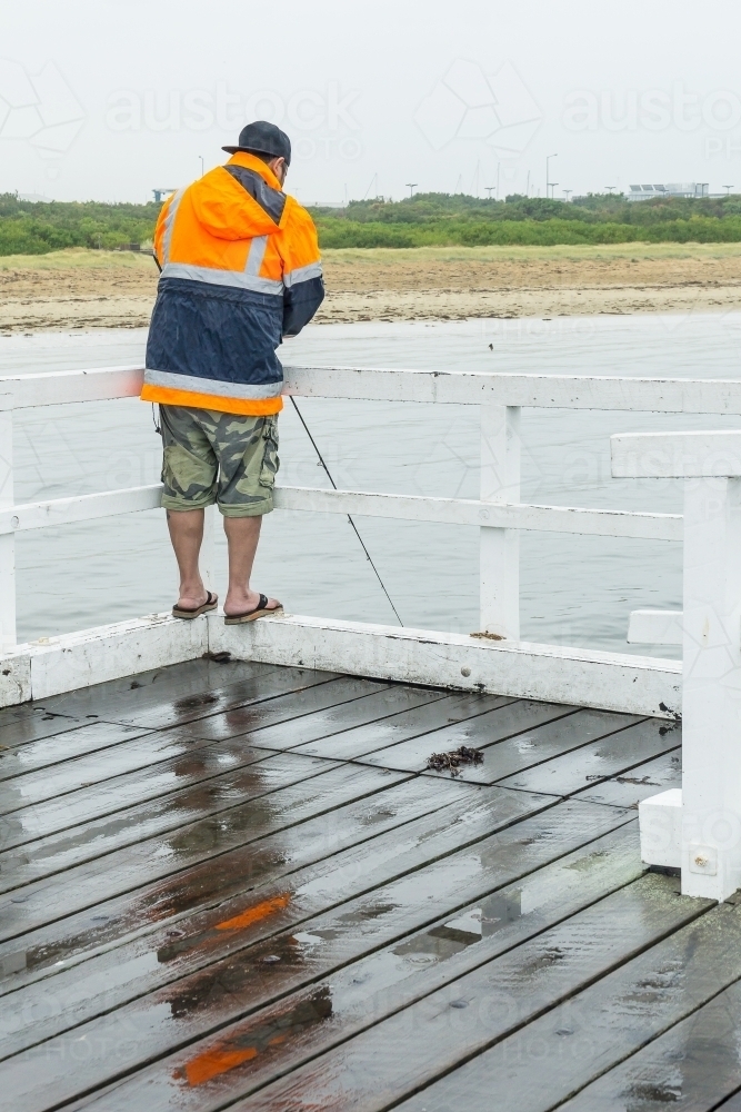 A man fishing over a jetty railing in wet weather - Australian Stock Image