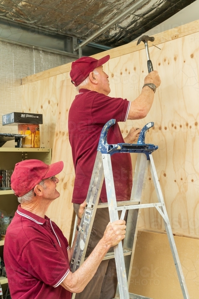 A man holding ladder while another man hammers against a wall in a Men's shed. - Australian Stock Image