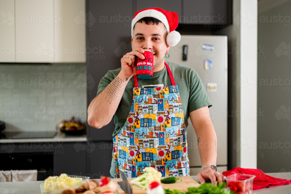 A man in a colourful apron enjoys a drink while cooking a festive lunch in his kitchen - Australian Stock Image