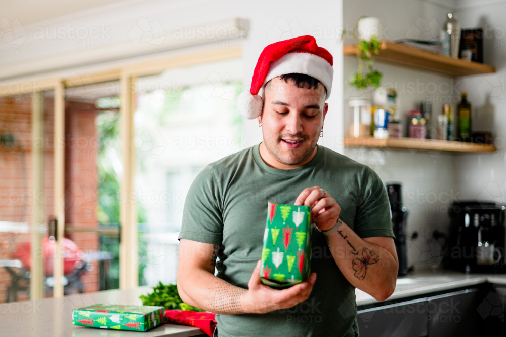 A man in a Santa hat excitedly unwraps a colourful present at home, celebrating Christmas - Australian Stock Image