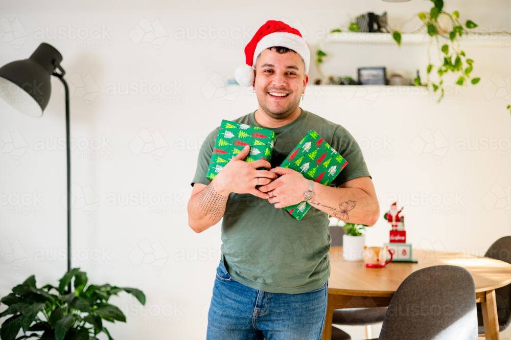 A man in a Santa hat smiles in a bright living room, holding Christmas gifts - Australian Stock Image