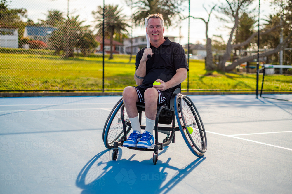 A man in a wheelchair holds a tennis ball, enjoying practice on a sunny day at a tennis court - Australian Stock Image