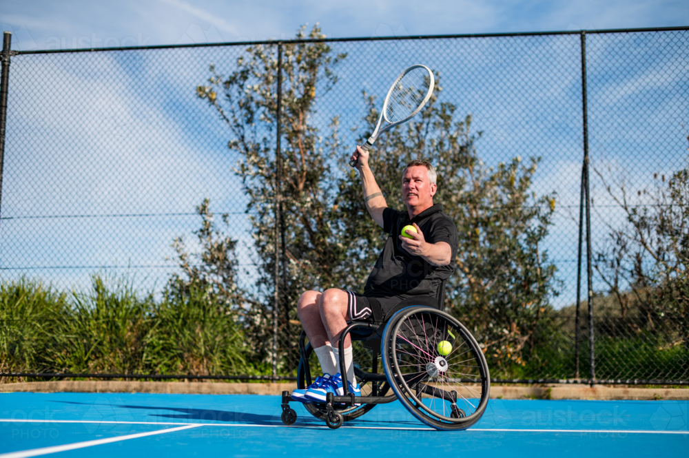 A man in a wheelchair is serving a tennis ball on an outdoor blue court on a sunny day - Australian Stock Image