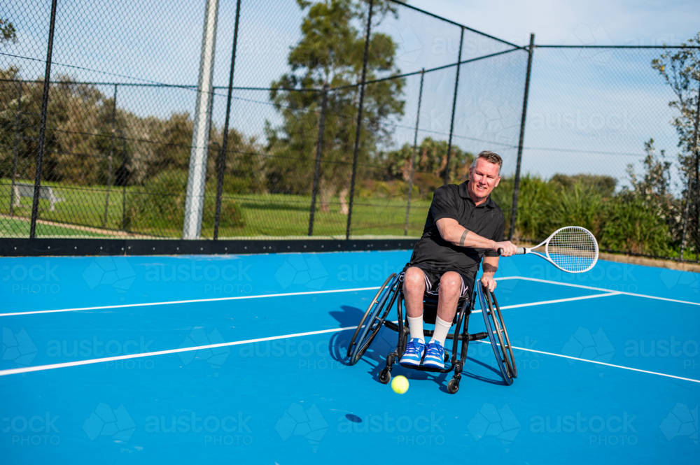 A man in a wheelchair plays tennis on a vibrant blue court in a sunny area - Australian Stock Image