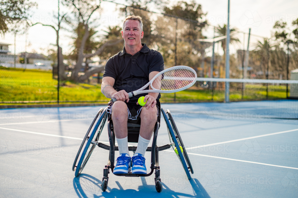 A man in a wheelchair poses with a tennis racquet and ball at a bright court - Australian Stock Image
