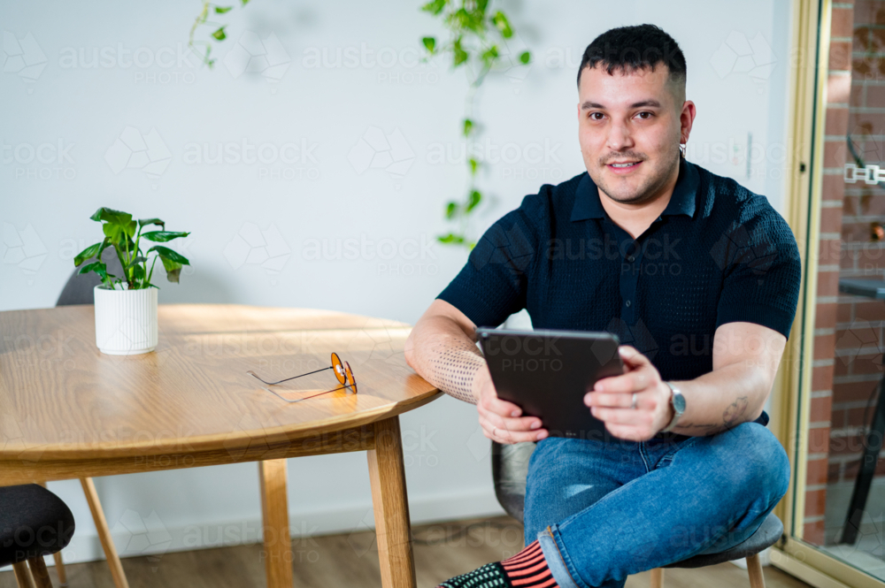 A man in his 30s enjoys reading on a tablet device while seated at a wooden table in a stylish home. - Australian Stock Image