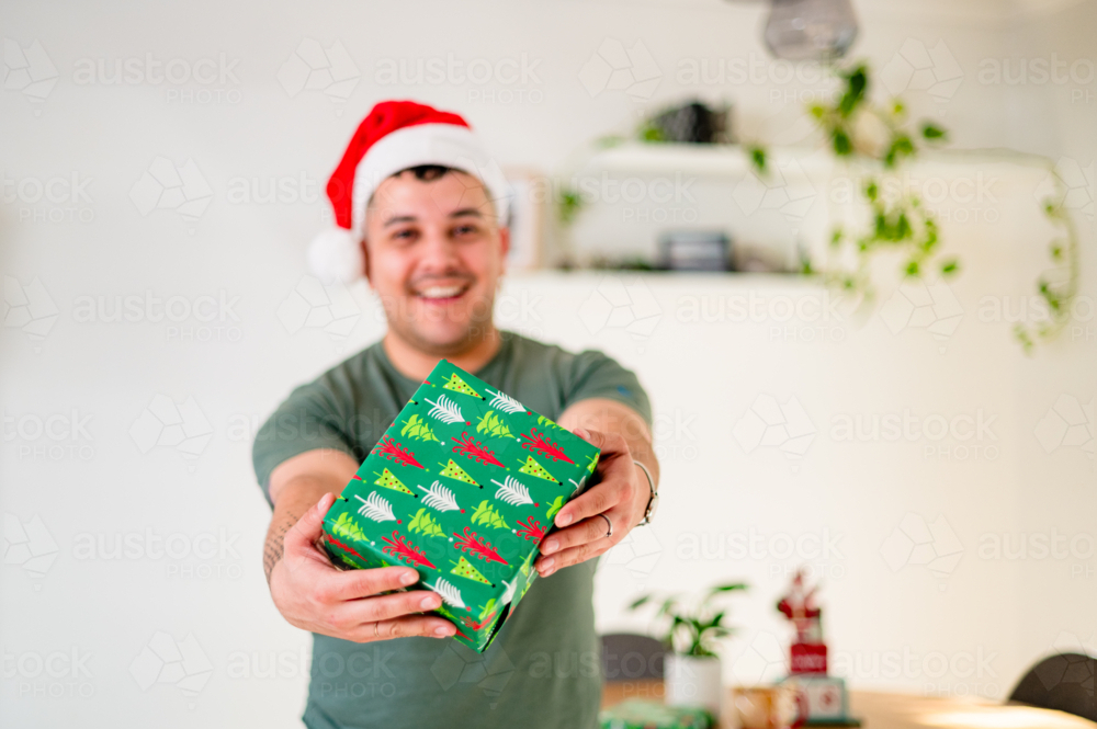 A man in his 30s in a Santa hat joyfully presents a gift while celebrating Christmas. - Australian Stock Image
