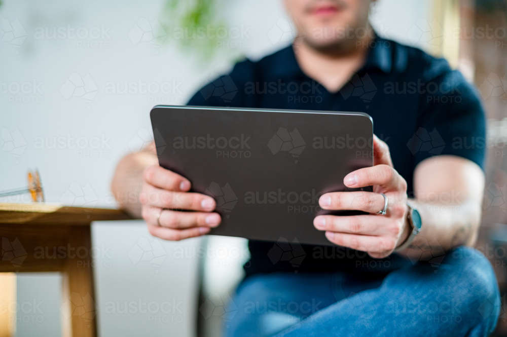 A man in his 30s sits relaxed at home, holding a tablet and engaging with digital content - Australian Stock Image