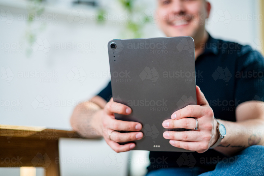 A man in his 30s smiles while holding a tablet at a wooden table in a cosy home setting - Australian Stock Image