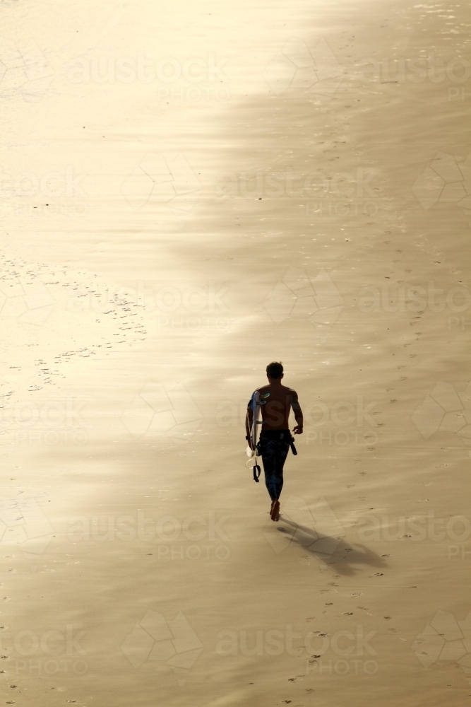 A man in his thirties jogging along beach with surfboard. - Australian Stock Image