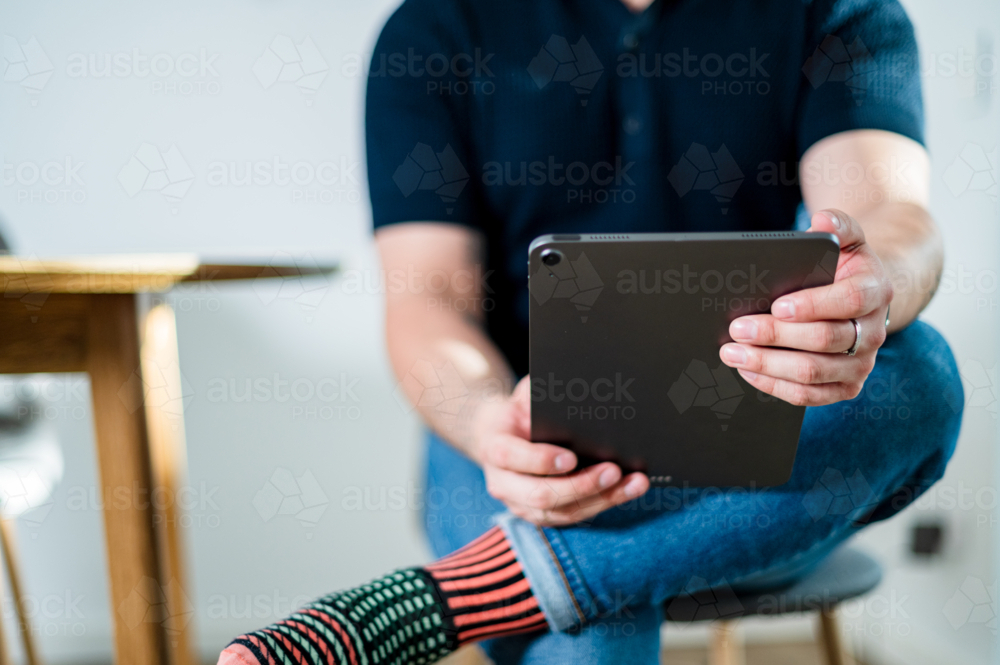 A man in his thirties sits at home, engaging with content on his tablet - Australian Stock Image