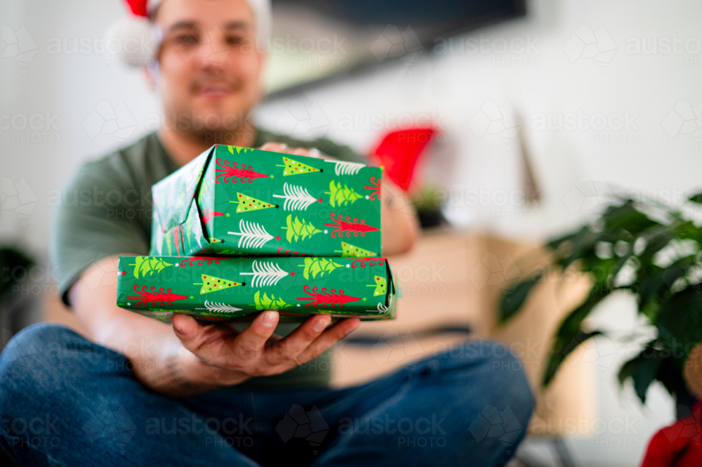 A man in his thirties smiles while holding wrapped gifts in a cosy, decorated space. - Australian Stock Image