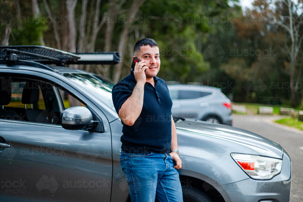 A man in his thirties stands outside his car, smiling as he chats on his phone. - Australian Stock Image