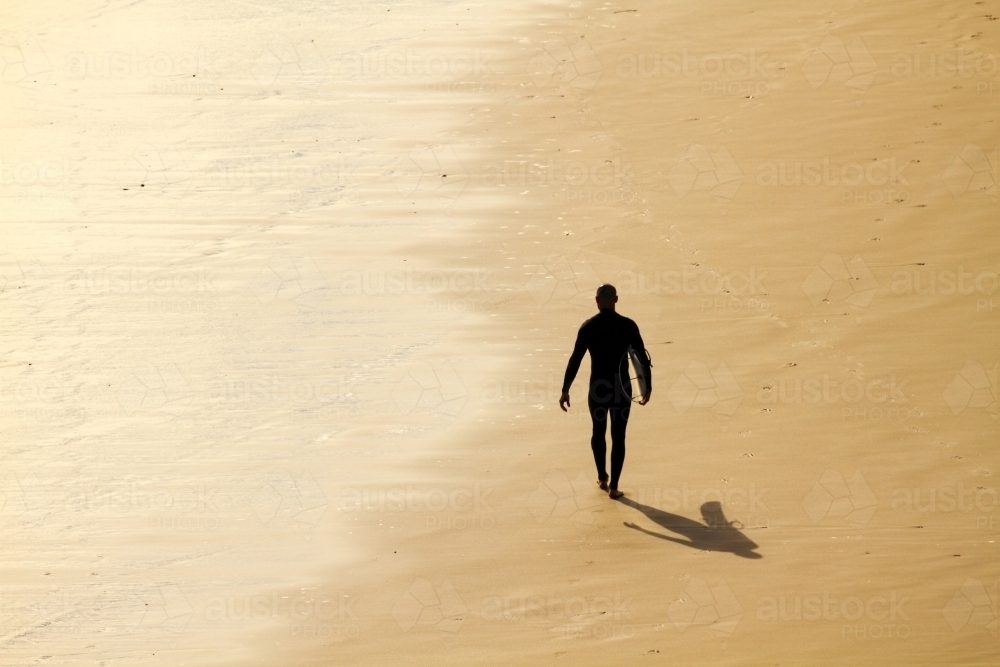 A man in his thirties walks along beach with surfboard. - Australian Stock Image