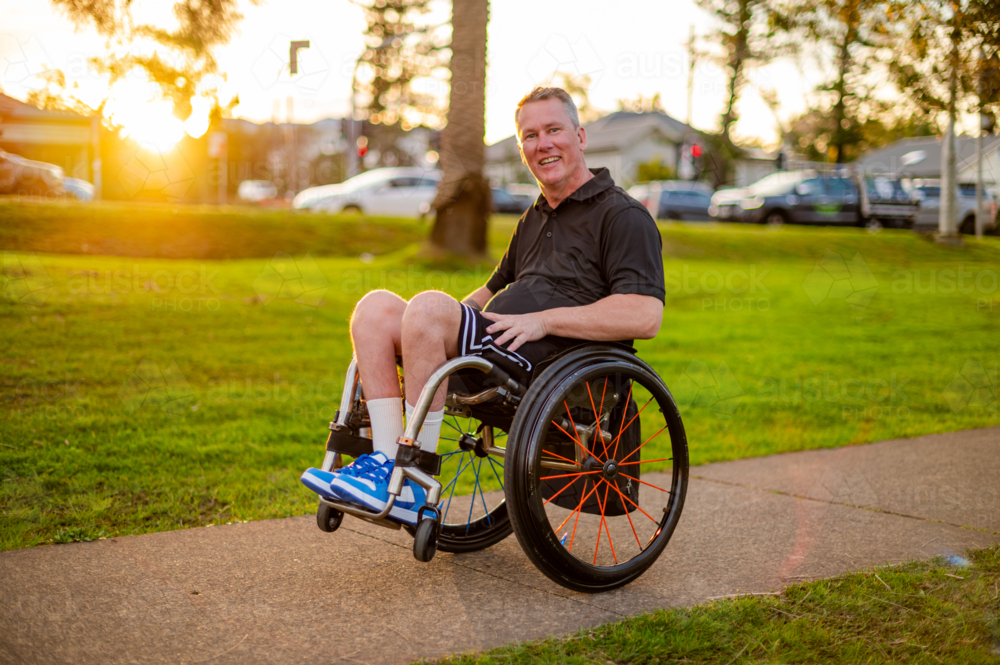 A man rides his wheelchair along a footpath while the sun sets in the background - Australian Stock Image