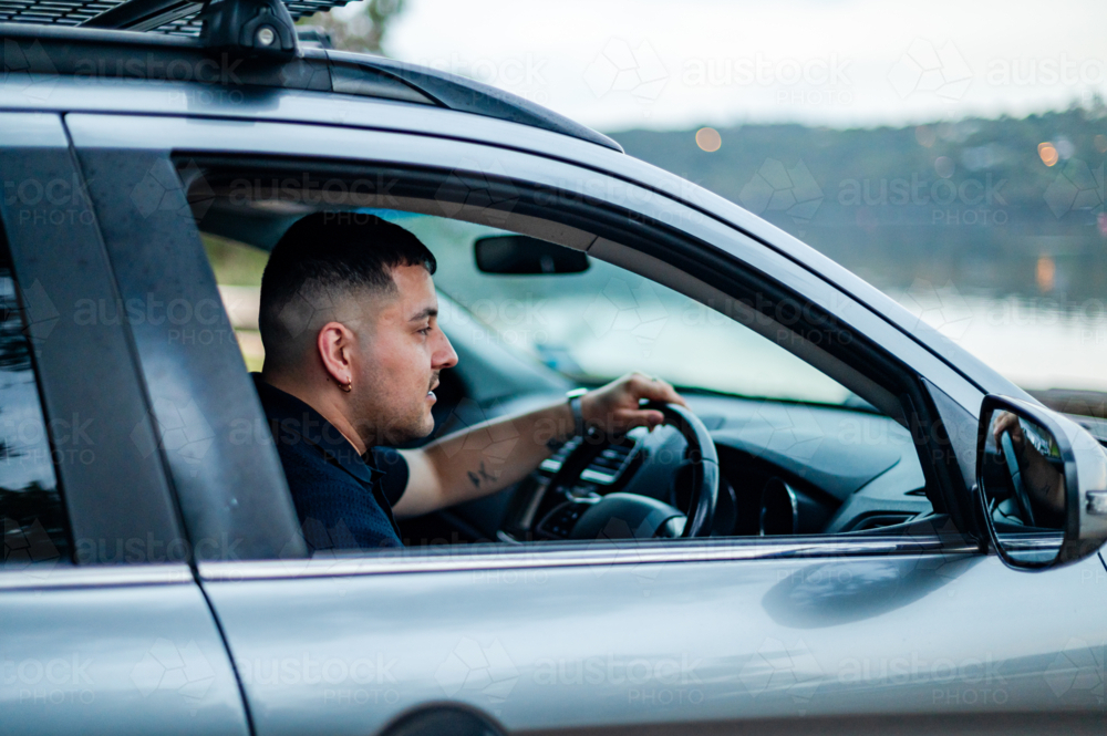 A man sits in a car, focused on the road beside a tranquil lake in a park during daylight. - Australian Stock Image