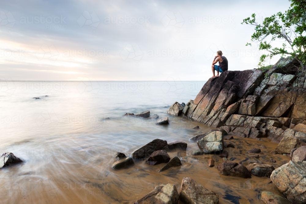 A man sits on a boulder in contemplation at a beautiful beach in a long exposure seascape scene. - Australian Stock Image