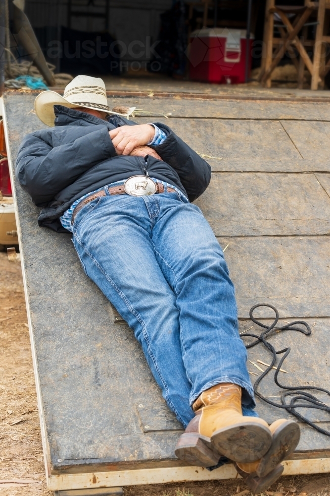 A man sleeping on ramp of a horse float with a hat over his face - Australian Stock Image