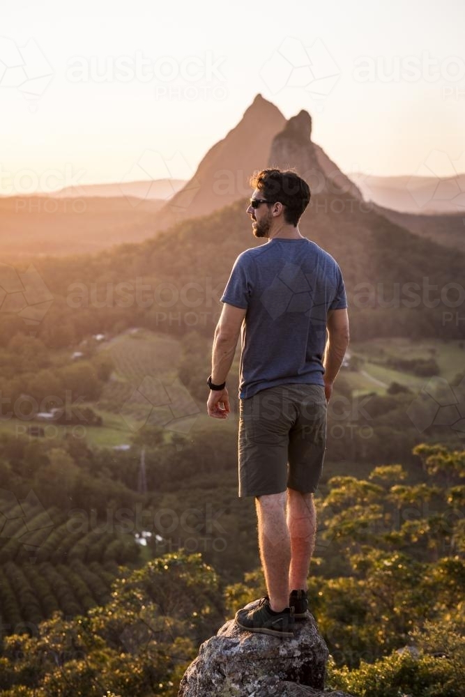 A man stands on a rock overlooking mountains - Australian Stock Image