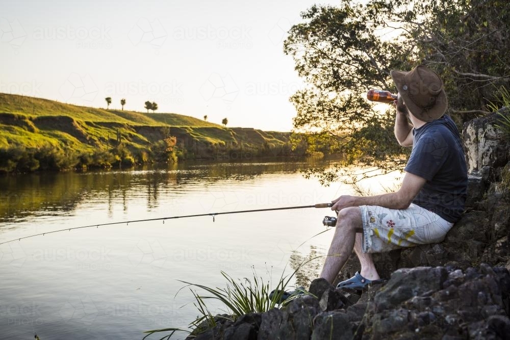 A man takes a drink whilst fishing by a river - Australian Stock Image