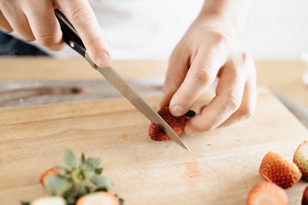 A man wearing a white t-shirt chopping up strawberries in a kitchen on a wooden board with a knife - Australian Stock Image