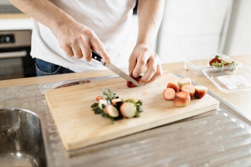A man wearing a white t-shirt chopping up strawberries in a kitchen on a wooden board with a knife - Australian Stock Image