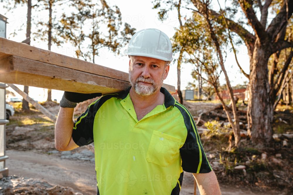 A man wearing hardhat carrying a stack of lumber on his shoulder at a construction site. - Australian Stock Image
