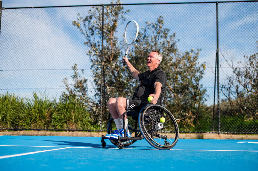 A mid aged man joyfully plays wheelchair tennis, racquet in hand, tossing tennis balls. - Australian Stock Image