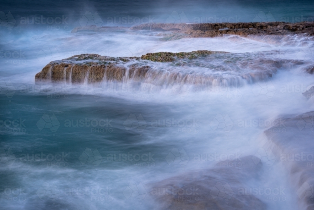 A moody, sombre long exposure of waves breaking over rocks - Australian Stock Image
