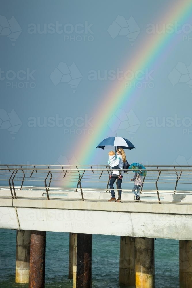 A mother and her children walk along a jetty under a rainbow - Australian Stock Image