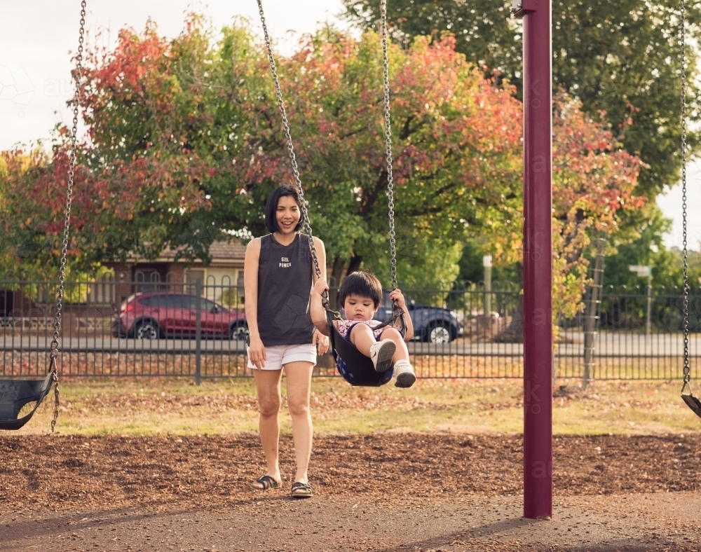 a mother pushing a toddler on a swing in autumn - Australian Stock Image