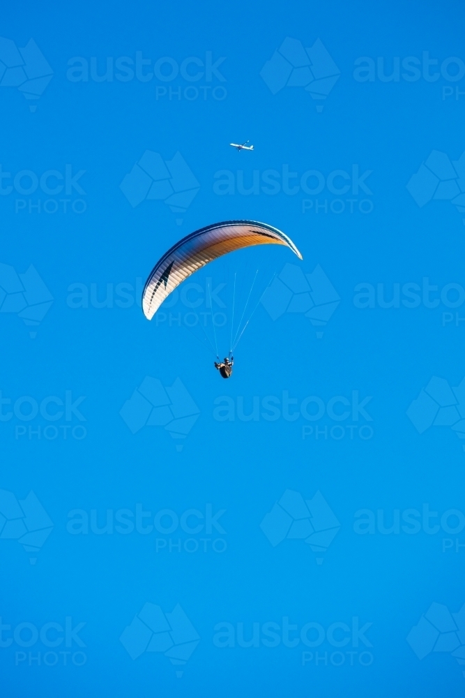 A paraglider floats in the clear blue sky with a distant aircraft above and in the background. - Australian Stock Image