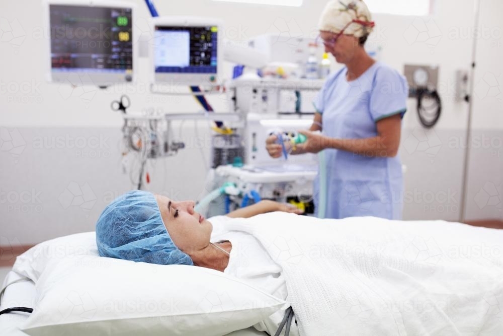 A patient lying on a bed in a hospital operating theatre with nurse and machines in background - Australian Stock Image