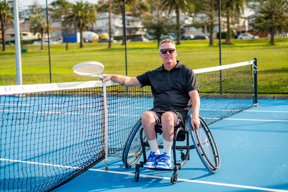 A person in a wheelchair with a tennis racquet smiles while enjoying tennis on a hard court - Australian Stock Image